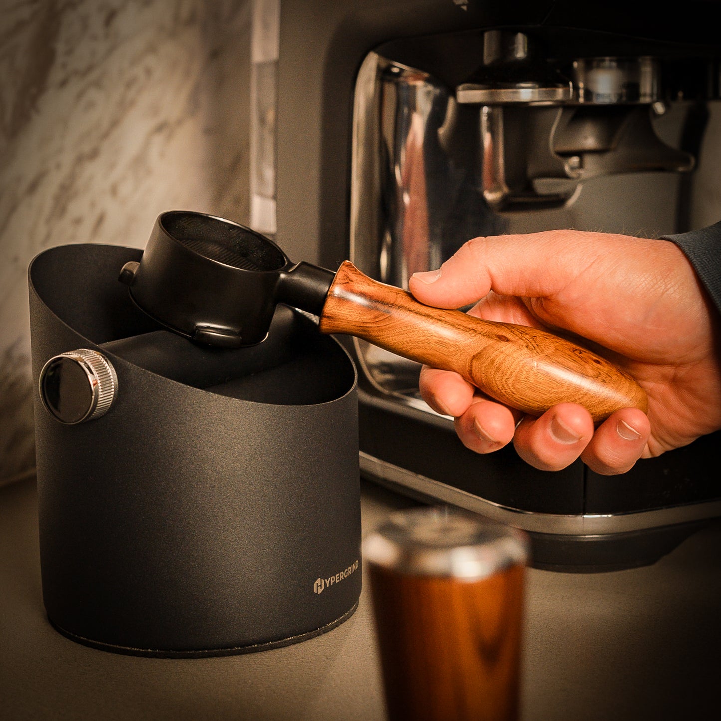 Hand holding a wooden-handled portafilter over a black coffee knock box with a coffee machine in the background.