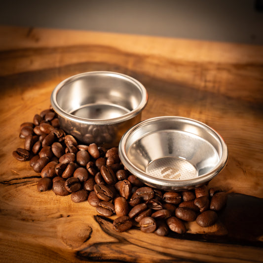 Two portafilter baskets on a wooden surface with coffee beans around them.
